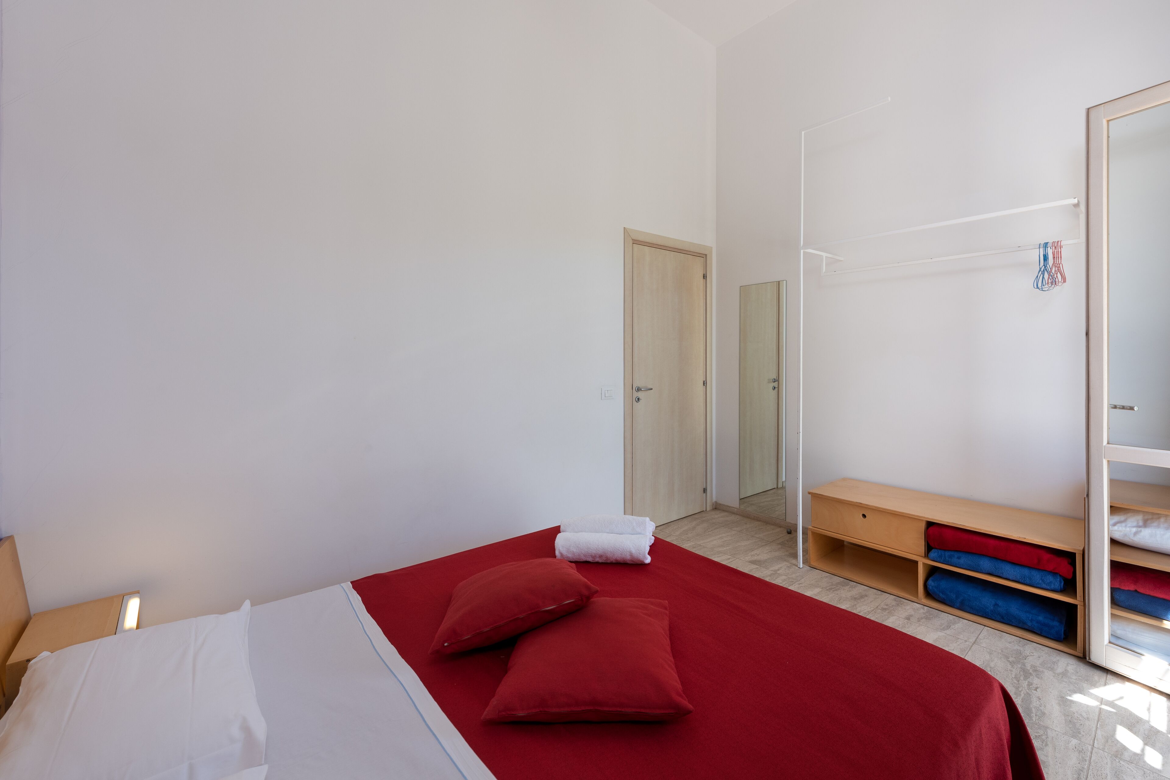 A bedroom with a bed, red and white pillows, a wooden bench, and a wardrobe with hanging clothes.