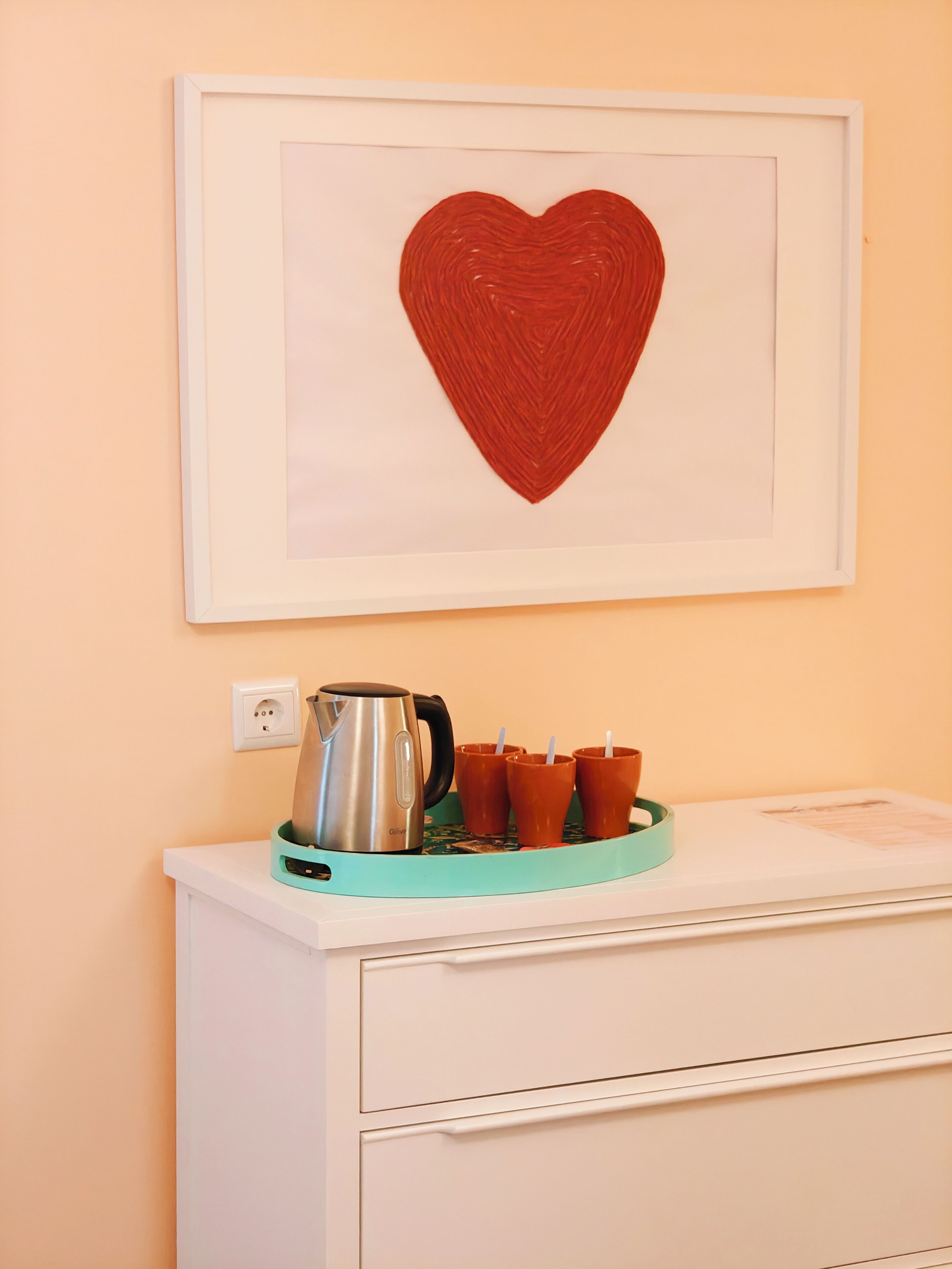 A framed red heart artwork on a wall above a white dresser with a teal tray holding a silver kettle and orange cups.
