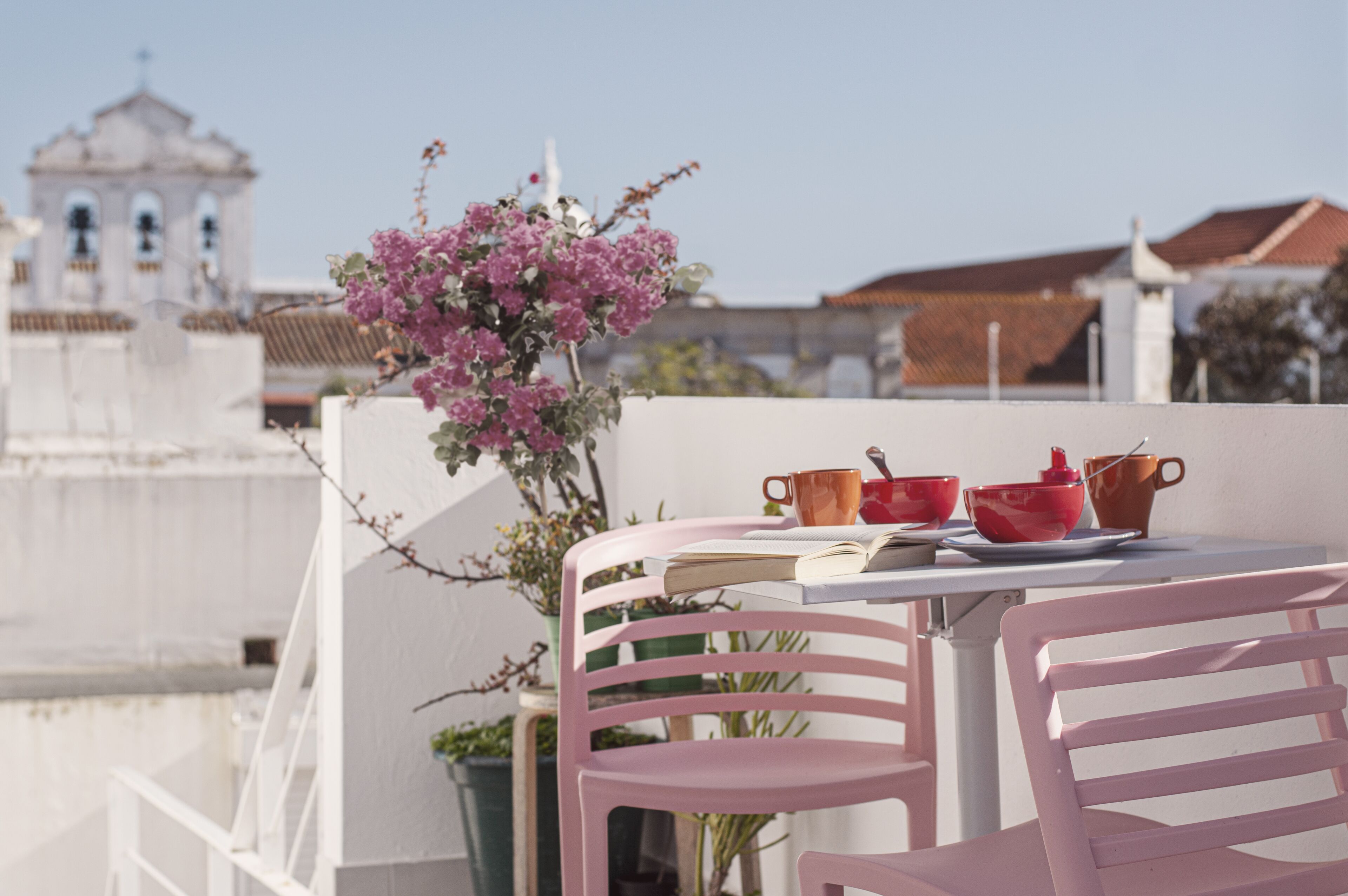 A balcony with a table set for tea, pink chairs, and a view of a church and buildings in the background.