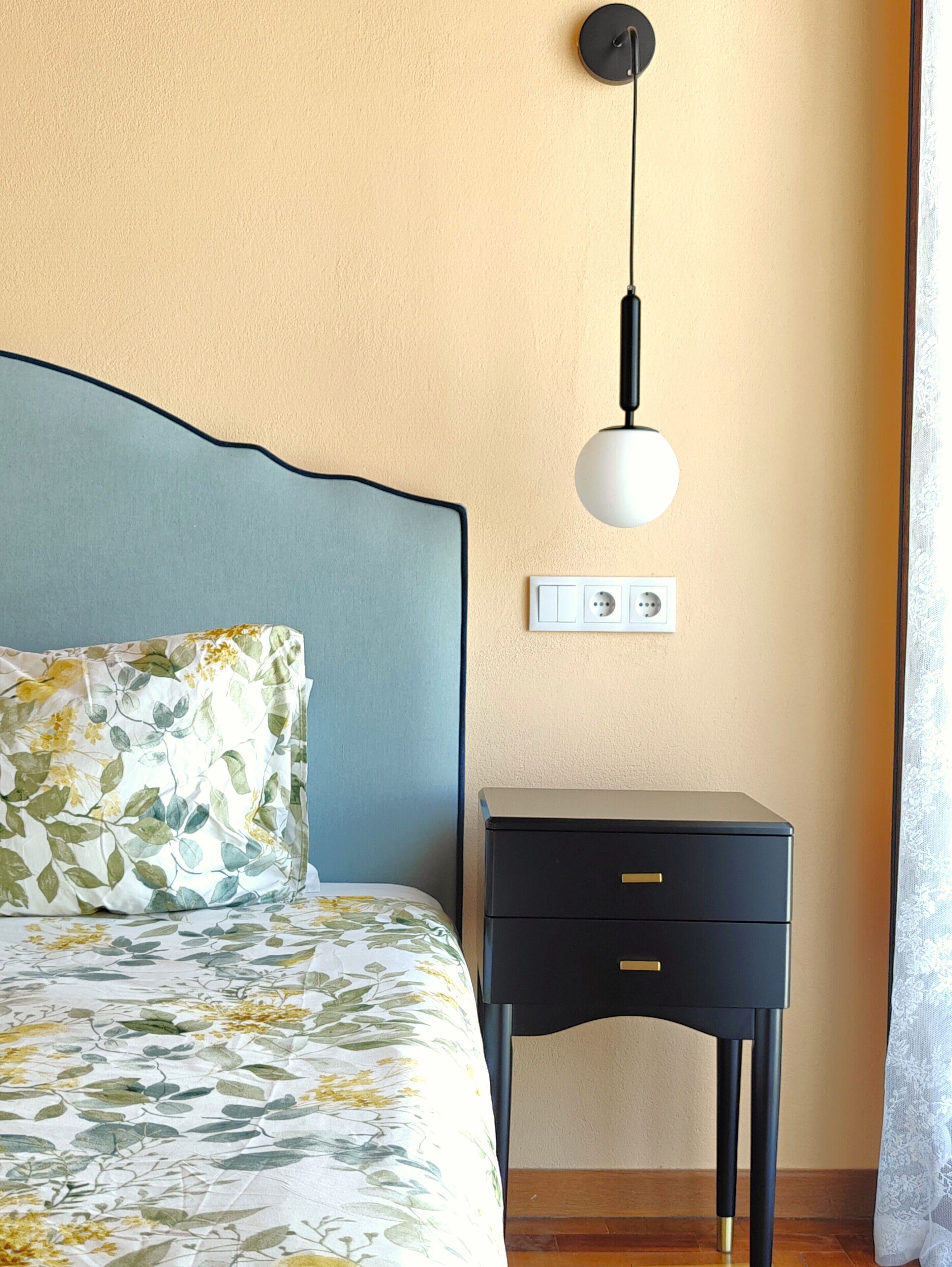 A neatly made bed with a floral-patterned pillow and a blue headboard, a bedside table with a drawer, and a wall-mounted light fixture.