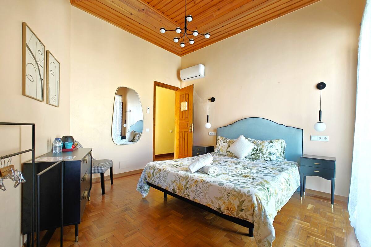 Un dormitorio con techo de madera, una cama con ropa de cama floral, un escritorio con silla, un espejo y un armario.