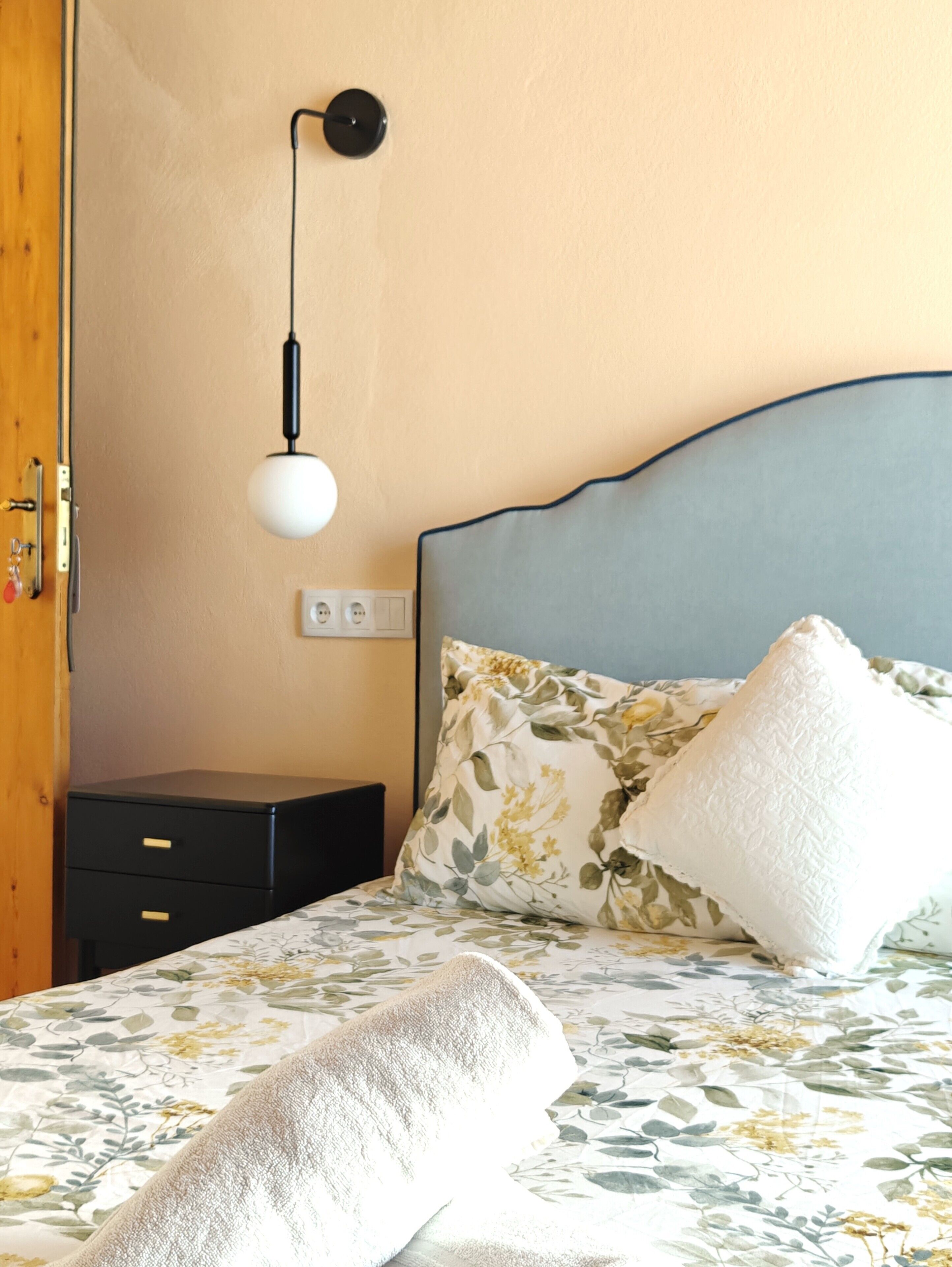 Ein ordentlich bezogenes Bett mit Blumenmuster, ein Nachttisch und eine Wandlampe.