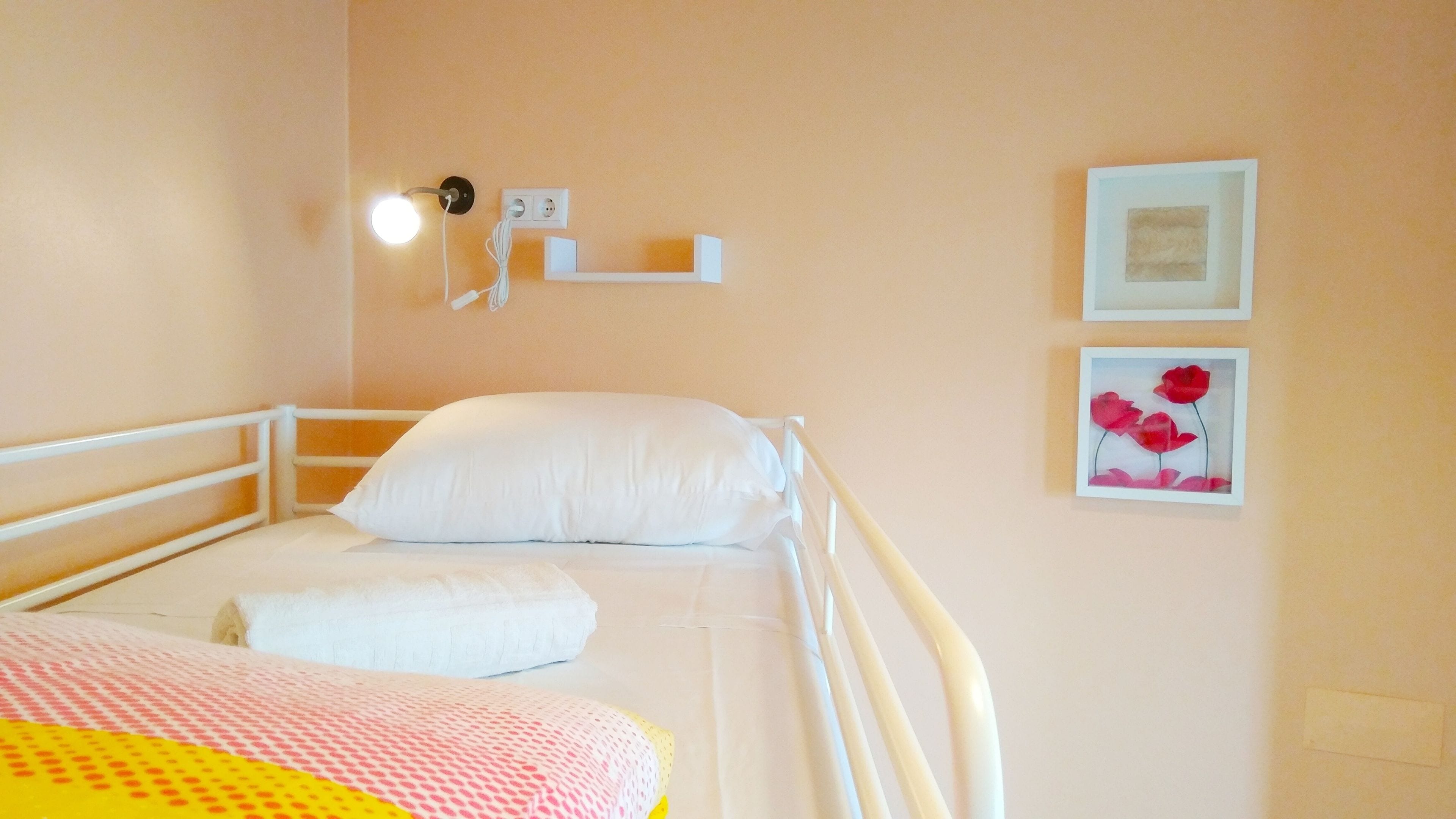 A single bed with a white headboard, a white pillow, and a white blanket. There are two wall-mounted light switches and a power outlet. Two framed pictures are on the wall.