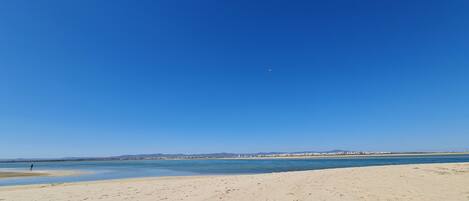 Een strand met voetafdrukken in het zand, een heldere blauwe lucht en een vliegtuig in de verte.
