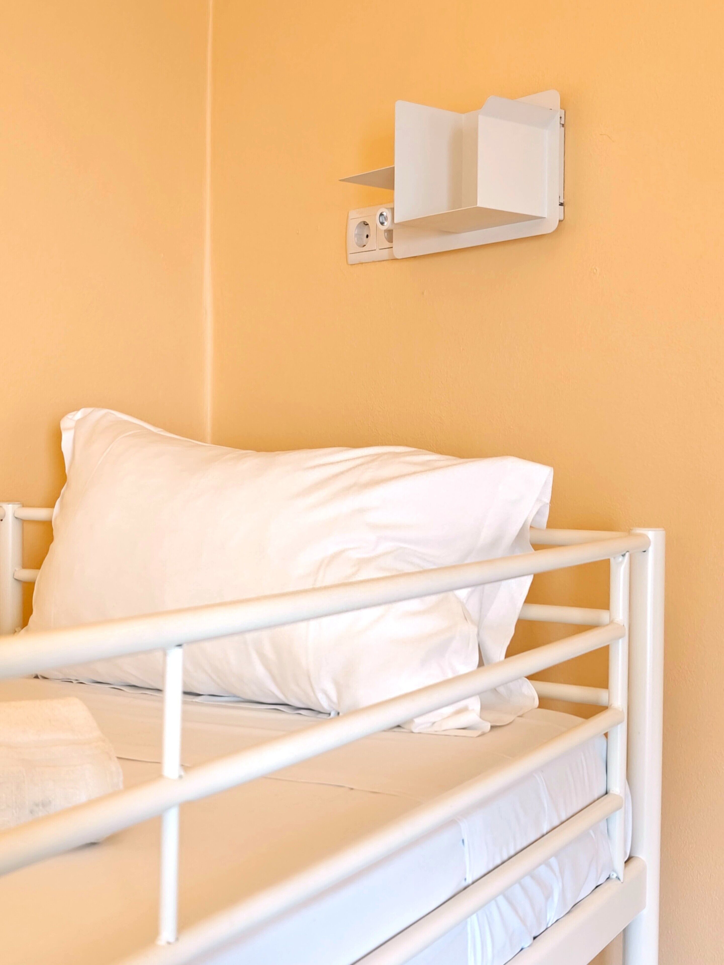 Una cama individual con ropa blanca y estructura metálica blanca, una lámpara fijada a la pared y una pared lisa de color amarillo.