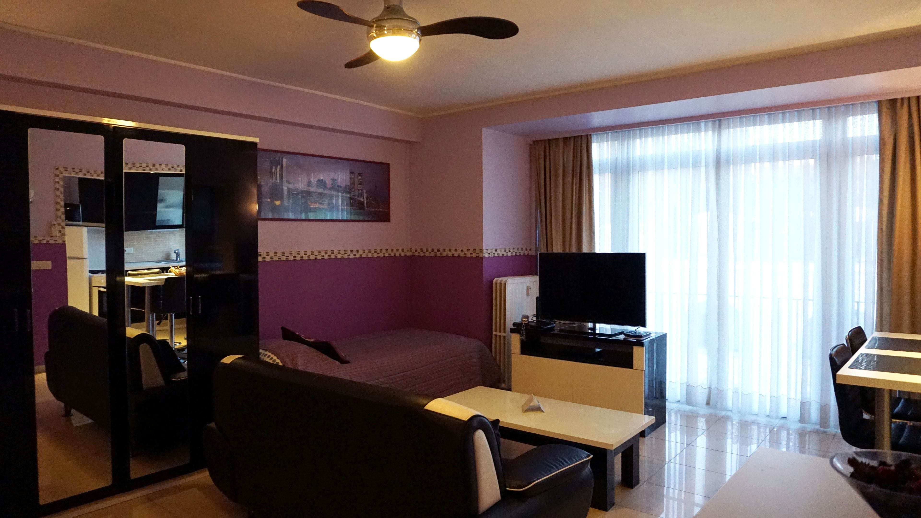 A hotel room with a bed, a desk with a computer, a TV, a sofa, and a large window with curtains.