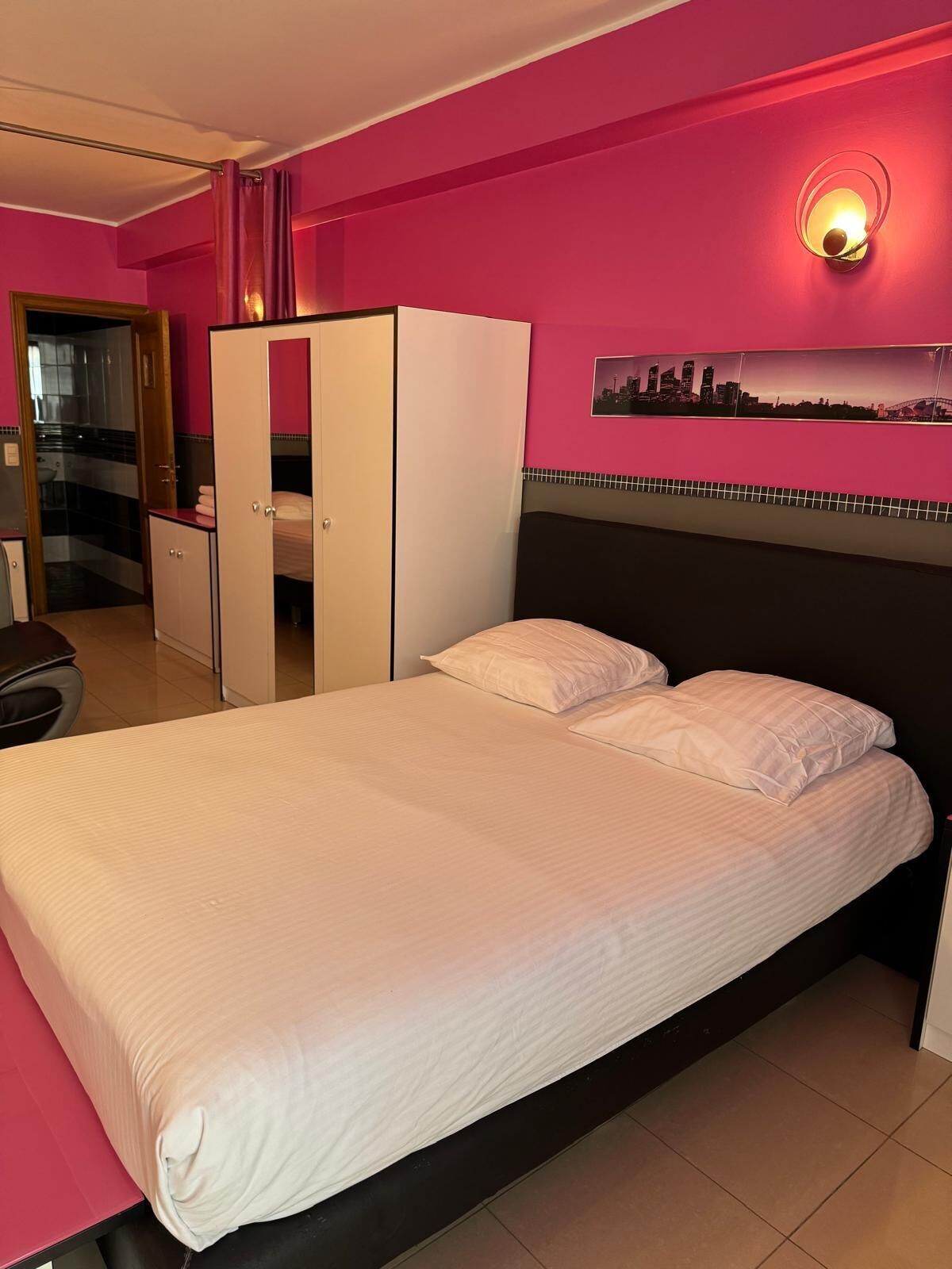 A hotel room with a bed, a wardrobe, a mirror, a sink, and a cityscape picture on the wall.