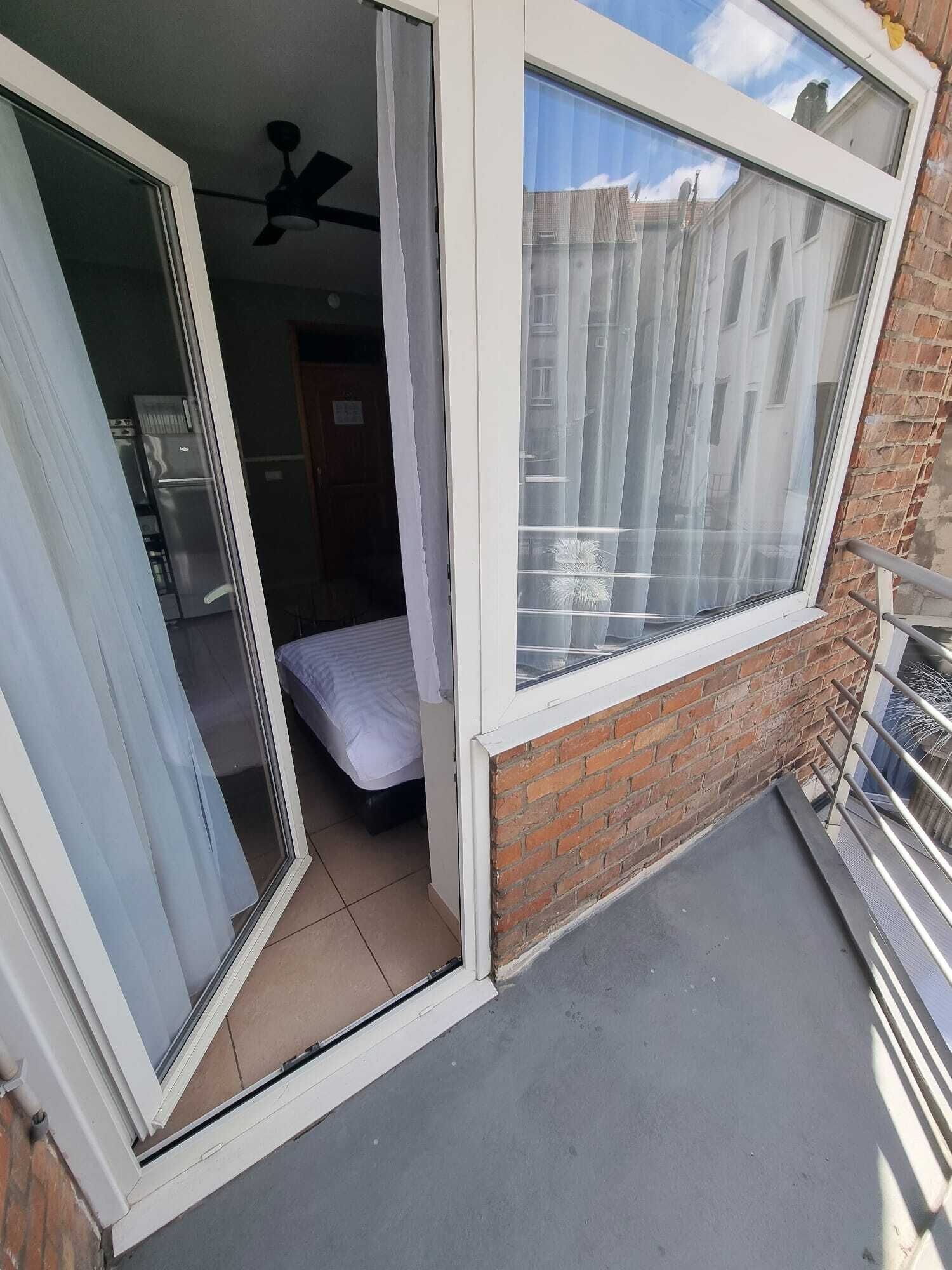 A balcony with a sliding glass door leading to a bedroom with a bed and a refrigerator.