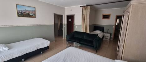 A hotel room with two single beds, a sofa, a ceiling fan, a painting, and a view of the city.