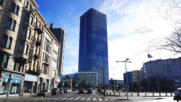 Un immeuble moderne de grande hauteur, doté d’une façade en verre, entouré de bâtiments plus anciens en briques, avec des devantures de magasins.
