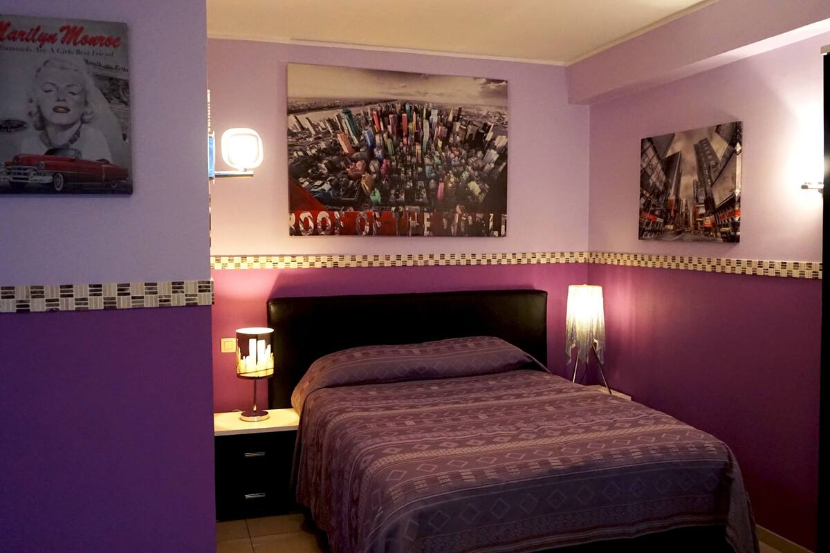 Un dormitorio con una cama, una mesita de noche, una lámpara y decoración en la pared, incluyendo un cartel de Marilyn Monroe y una pintura de un paisaje urbano.