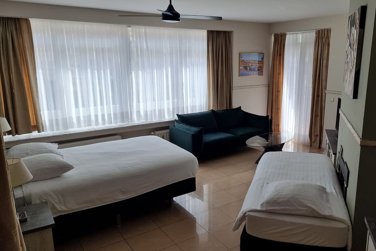 Habitación de hotel con dos camas individuales, un sofá verde oscuro, una mesita redonda y un ventilador de techo.