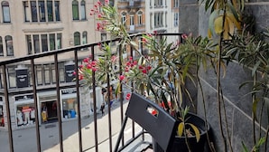 Un balcon avec une chaise et des plantes en pot, donnant sur une rue bordée d’immeubles et de boutiques.