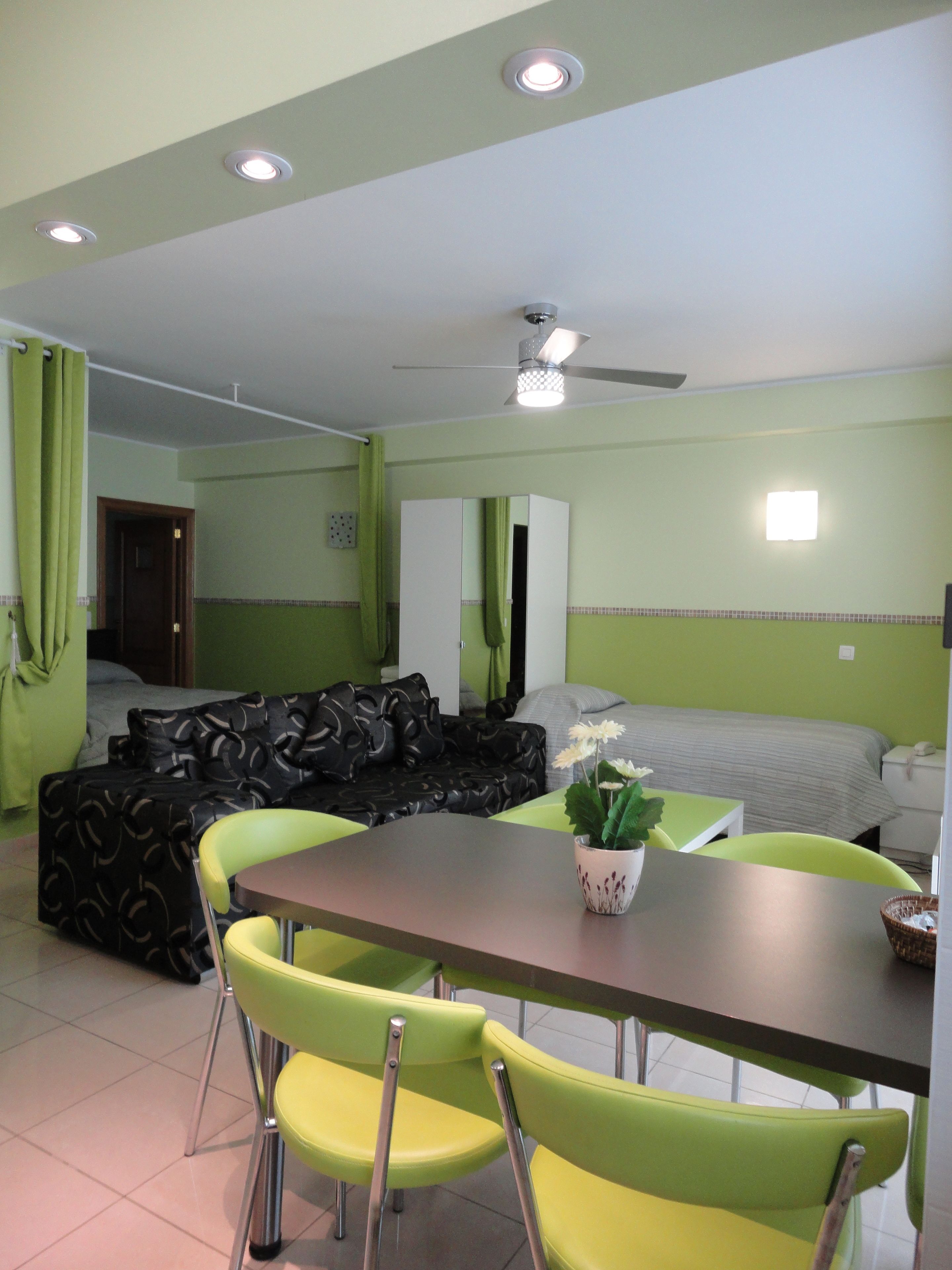 A room with a dining table, chairs, a sofa, a bed, and a ceiling fan.