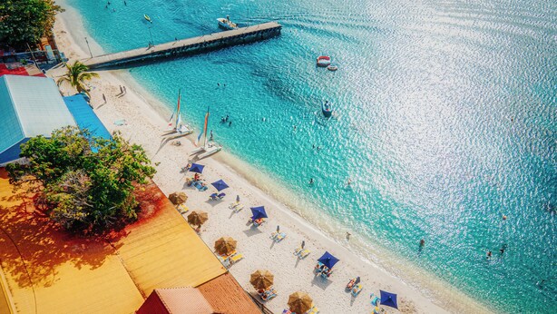 Een strand met helder turquoise water, een houten steiger en strandparasols.