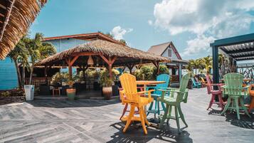 Un patio al aire libre con sillas y mesas de colores, una estructura de techo de paja y plantas en macetas.
