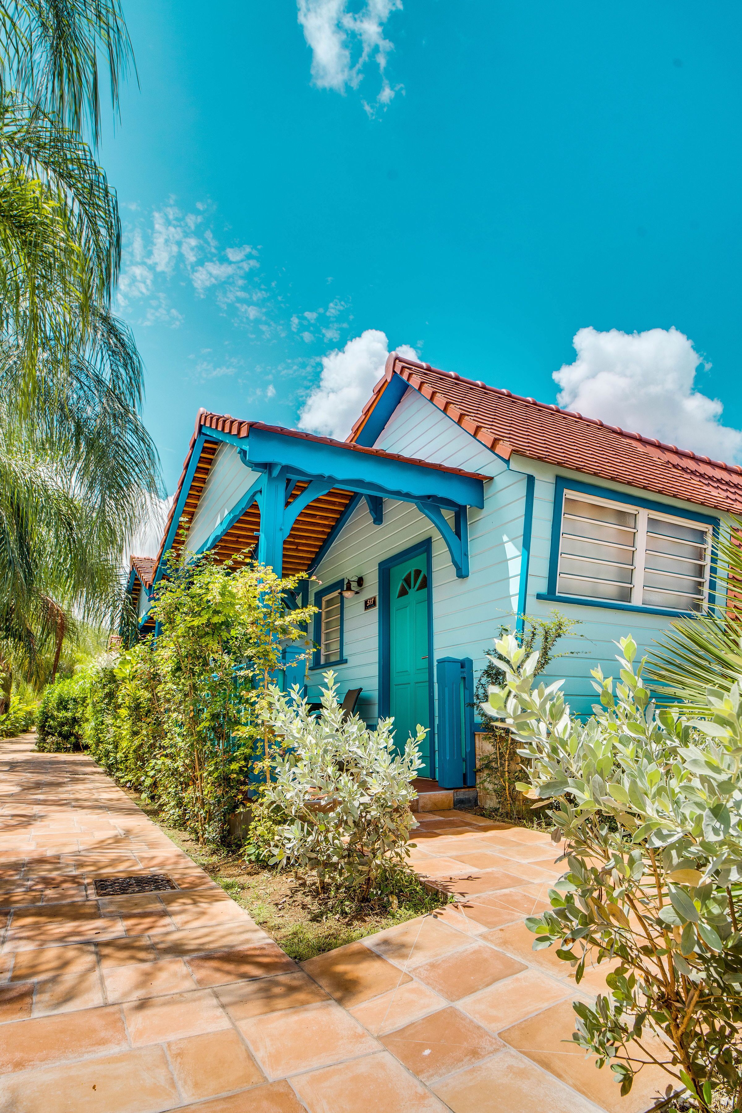 Una casa con fachada azul y techo rojo, rodeada de abundante vegetación y un cielo azul despejado.