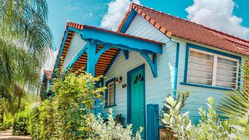 Una casa con fachada azul y techo rojo, rodeada de abundante vegetación y un cielo azul despejado.