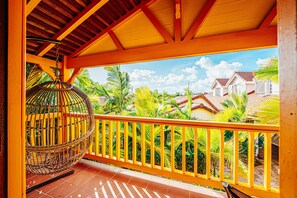 A balcony with a wicker chair, yellow railing, and a view of houses and palm trees. - Hôtel Bambou & Spa (Trois-Ilets)