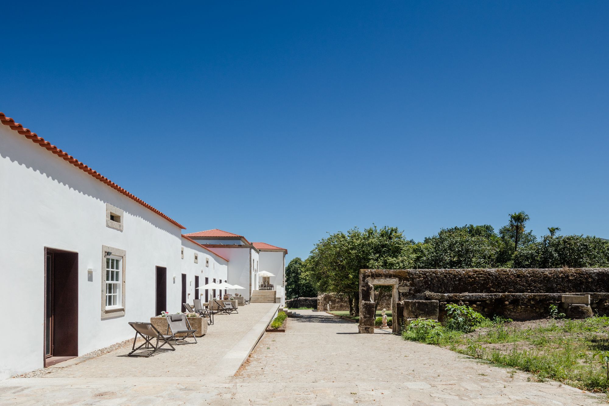 A white building with red-tiled roofs, a paved courtyard, outdoor seating, and a stone wall with vegetation.