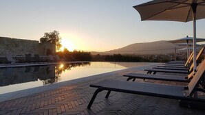 Un espace piscine extérieur paisible, doté de chaises longues et de parasols, entouré d’un mur de pierre et offrant une vue sur une étendue d’eau calme, avec un coucher de soleil en arrière-plan.