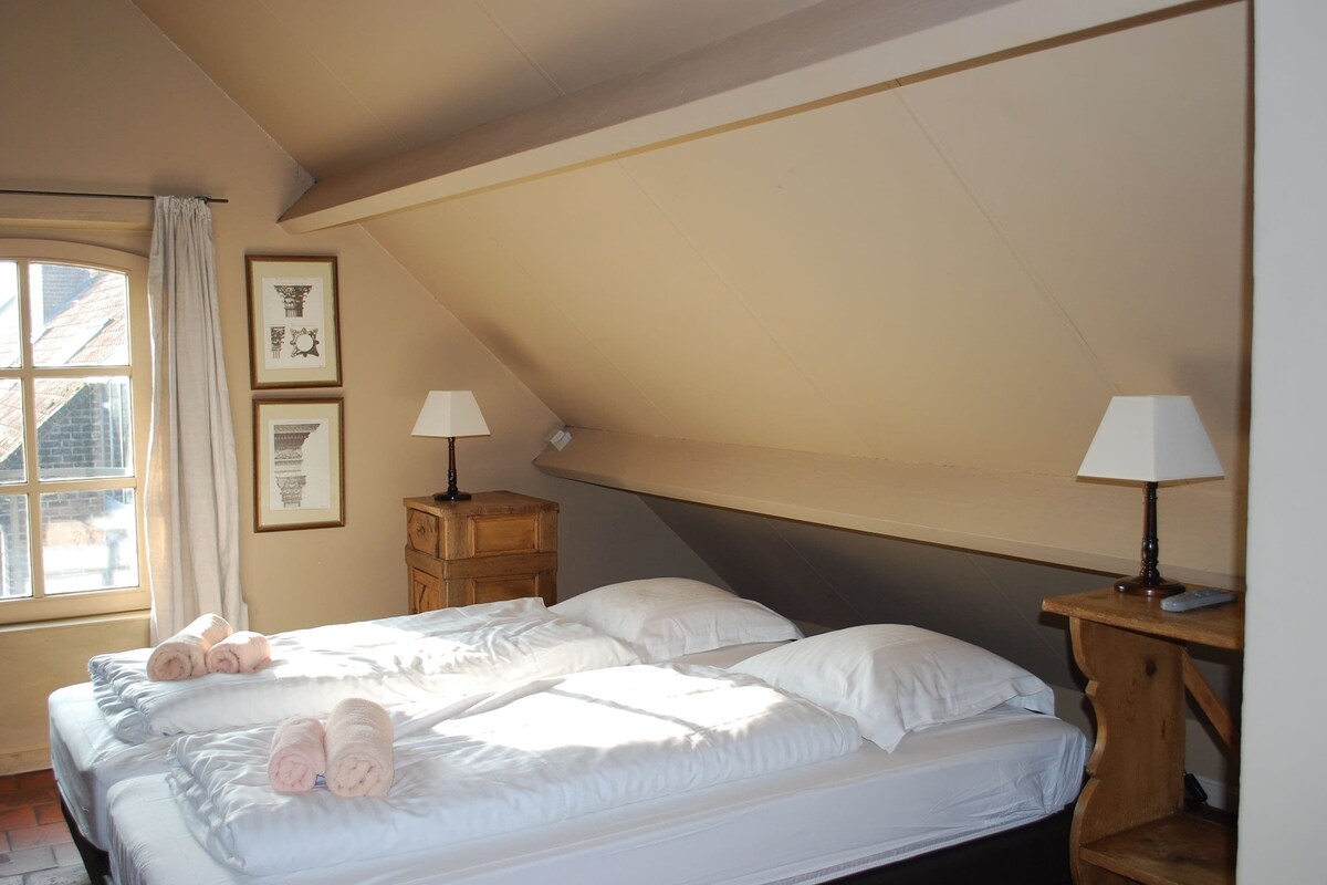 A bedroom with a bed, two bedside tables, a window with curtains, and framed pictures on the wall.