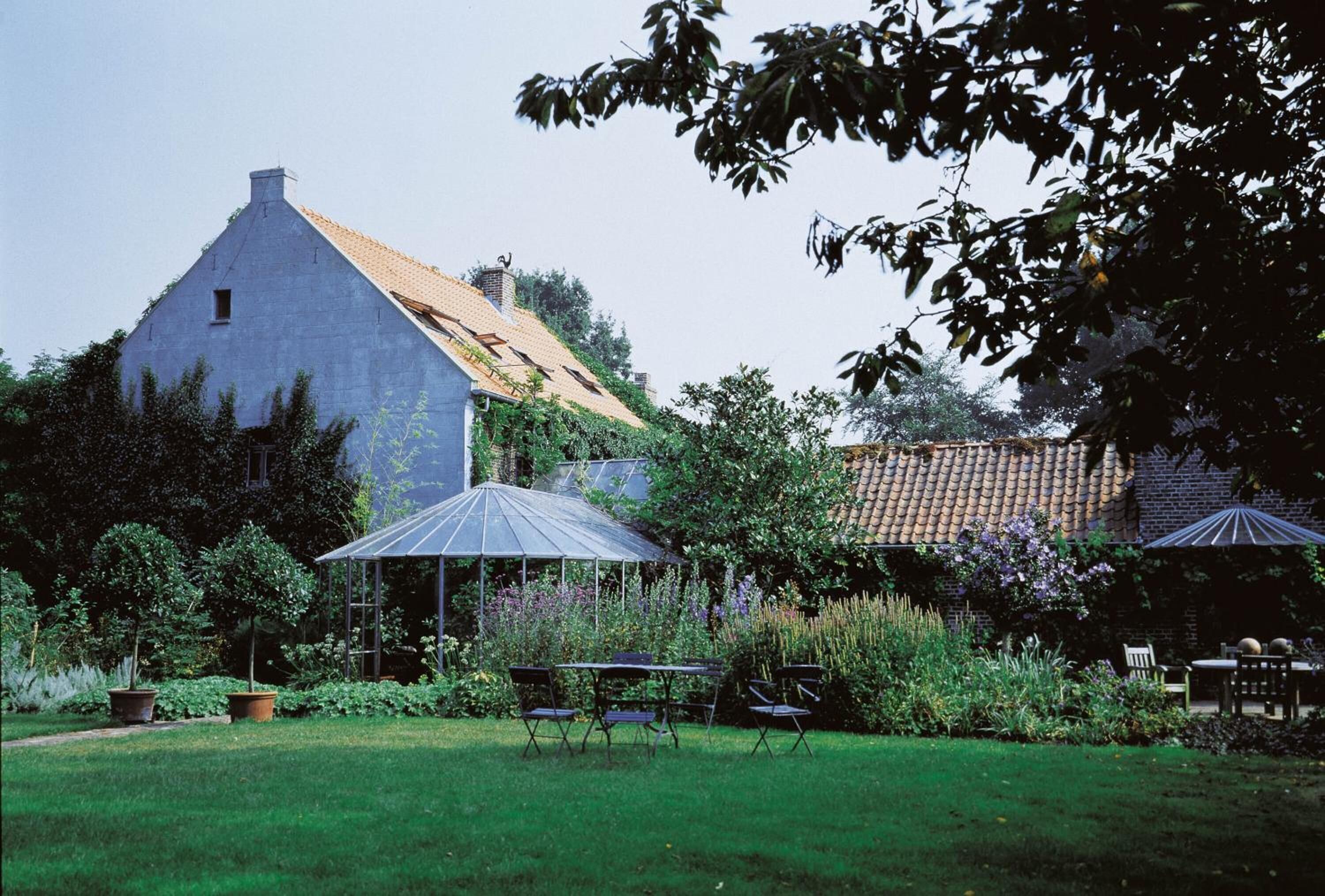 Een groot gebouw met een schuin dak, omringd door een verzorgde tuin met een prieel, buitenmeubels en potplanten.