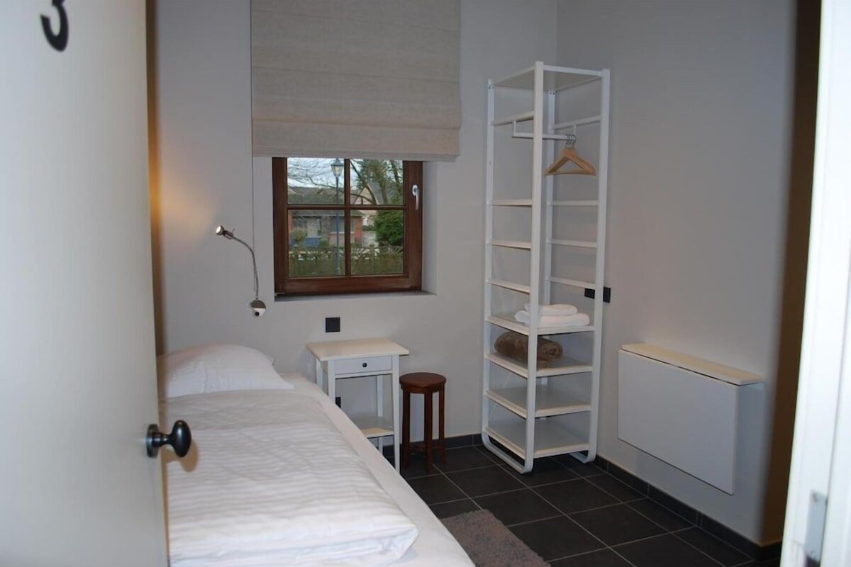 A bedroom with a bed, a bedside table, a lamp, a window, a radiator, and a white shelving unit.