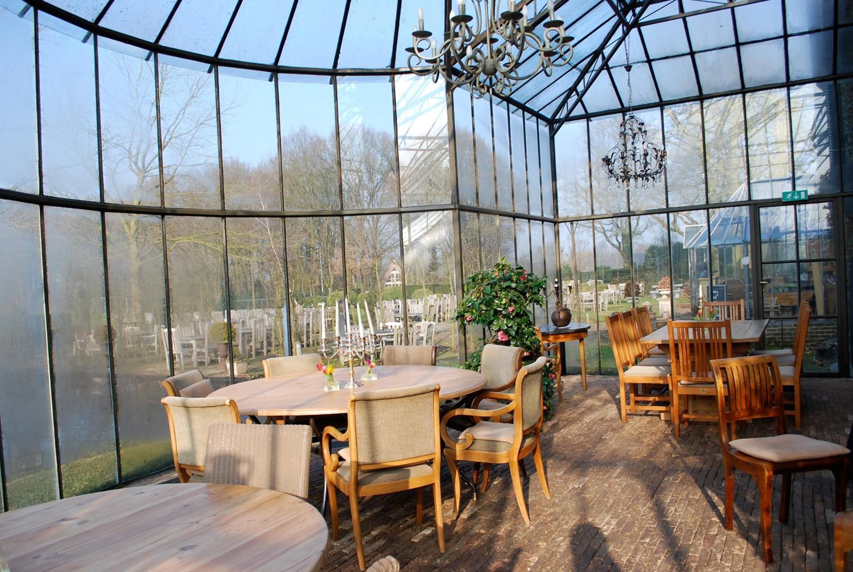 Een kas met glazen dak, houten tafels en stoelen klaar voor dineren.