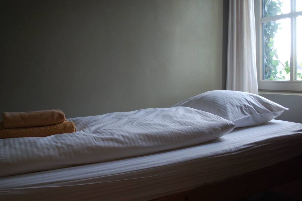 A bed with white bedding and a brown towel, a window with curtains, and a view of greenery outside.