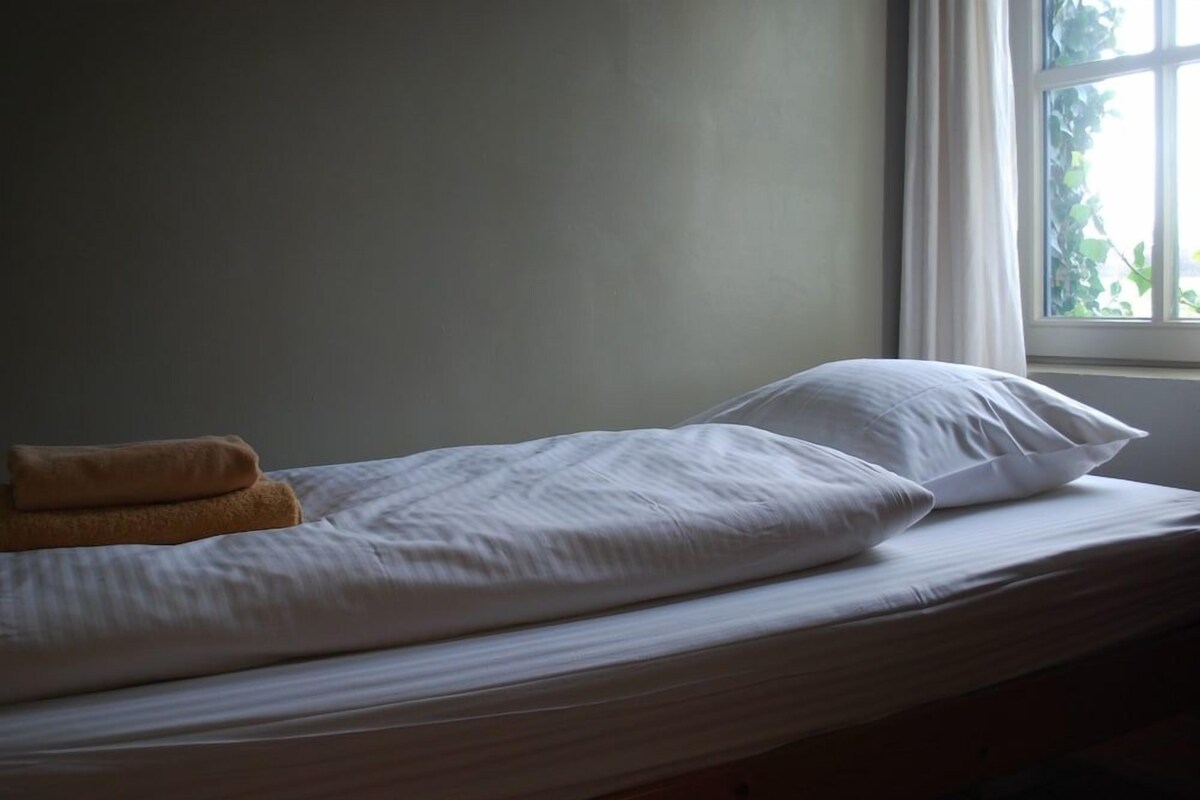 A single bed with white bedding and a brown towel, positioned next to a window with a view of greenery.