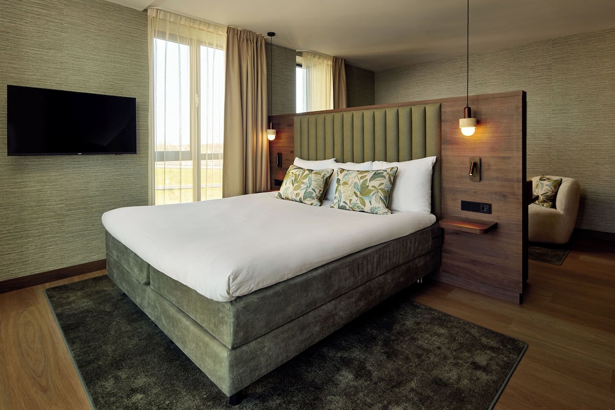 A modern hotel room with a large bed, a flat-screen TV mounted on the wall, a wooden headboard, and a small seating area with a chair.