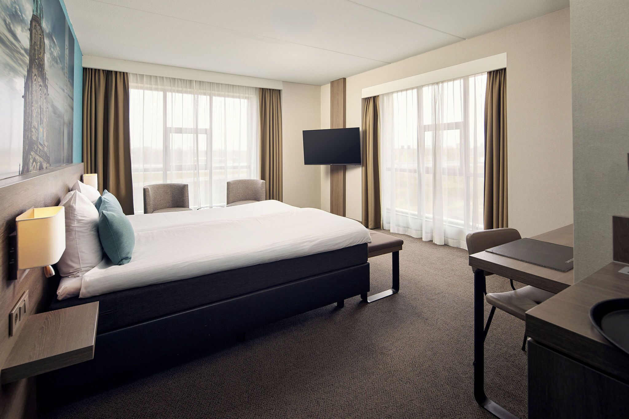 Habitación de hotel con cama, escritorio, silla, televisor y ventanas amplias.