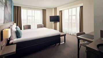 Habitación de hotel con cama, escritorio, silla, televisor y ventanas amplias.