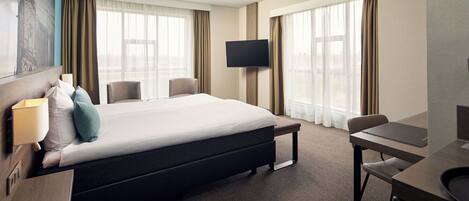 Habitación de hotel con cama, escritorio, silla, televisor y ventanas amplias.