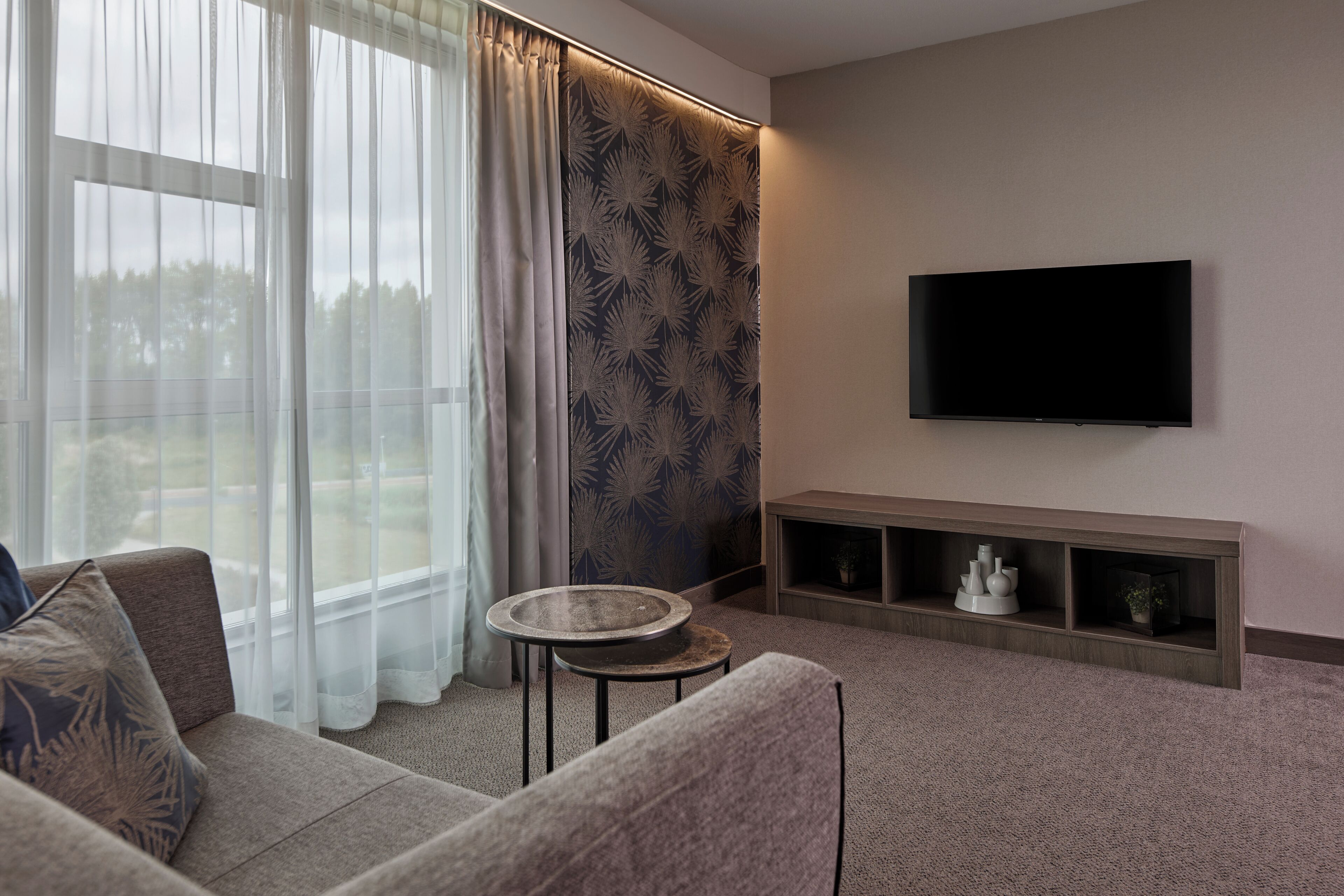 A living room with a sofa, a small round table, a flat-screen TV mounted on the wall, and a patterned curtain.
