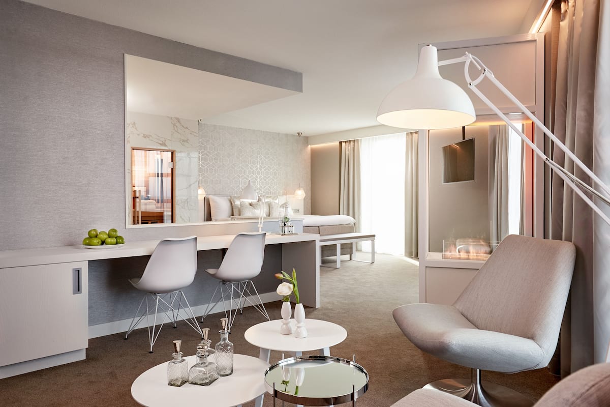 A modern hotel room with a dining area, a sitting area with a lamp, and a view of the city.