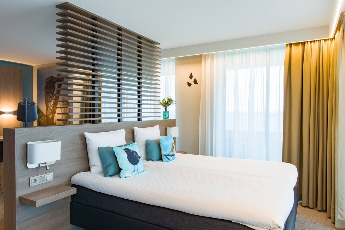 A modern hotel room with a large bed, a built-in desk, and a window with curtains.