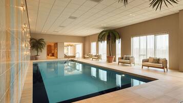 An indoor swimming pool with tiled walls, a ceiling with recessed lighting, and seating areas with potted plants.
