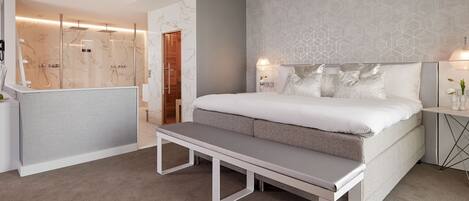 A modern bedroom with a large bed, a bench, a bedside table, and a shower area.