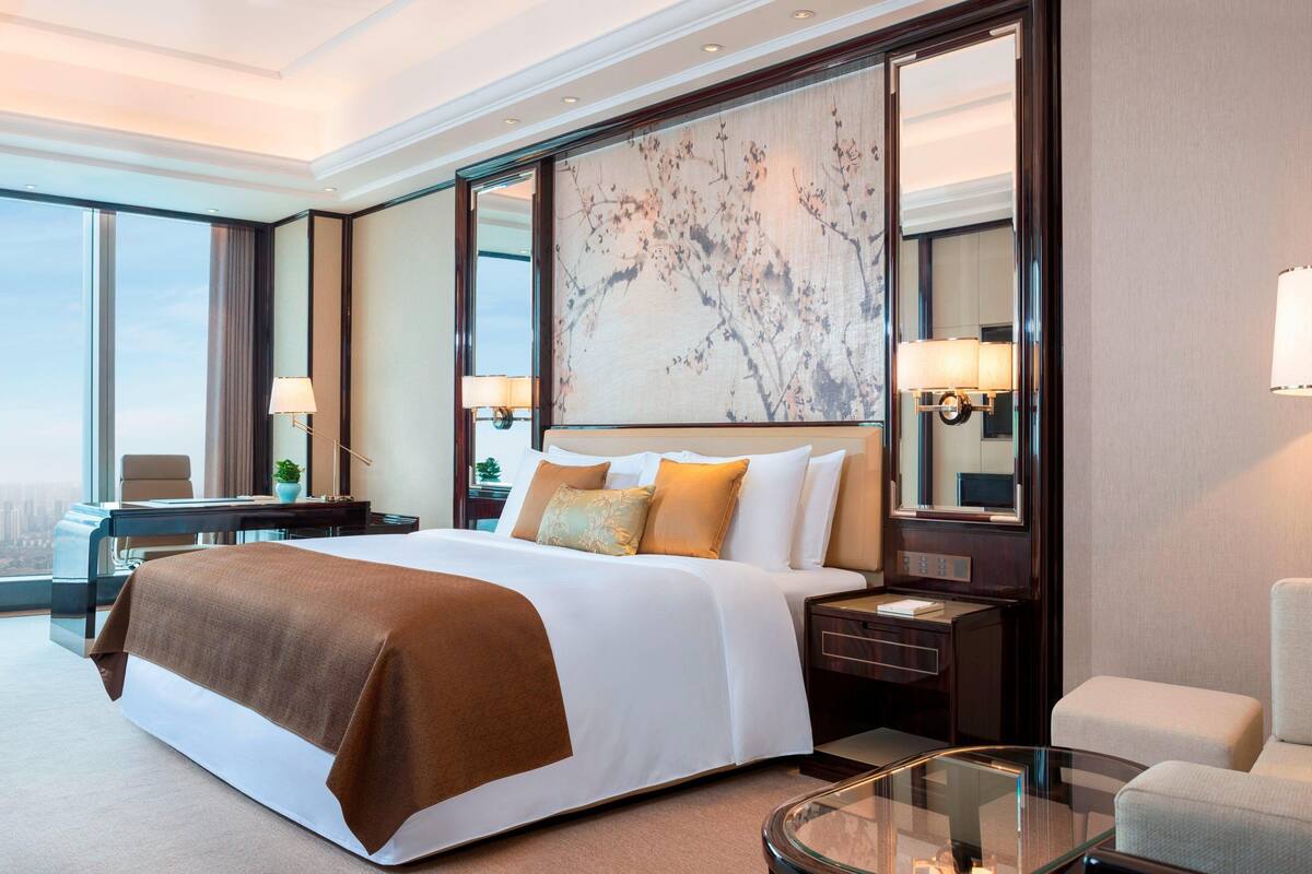 Ein Hotelzimmer mit einem großen Bett, einem Glastisch, einem Schreibtisch mit Lampe und Blick auf die Stadt.
