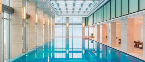 A modern indoor swimming pool with clear glass walls, a skylight ceiling, and a view of the cityscape.