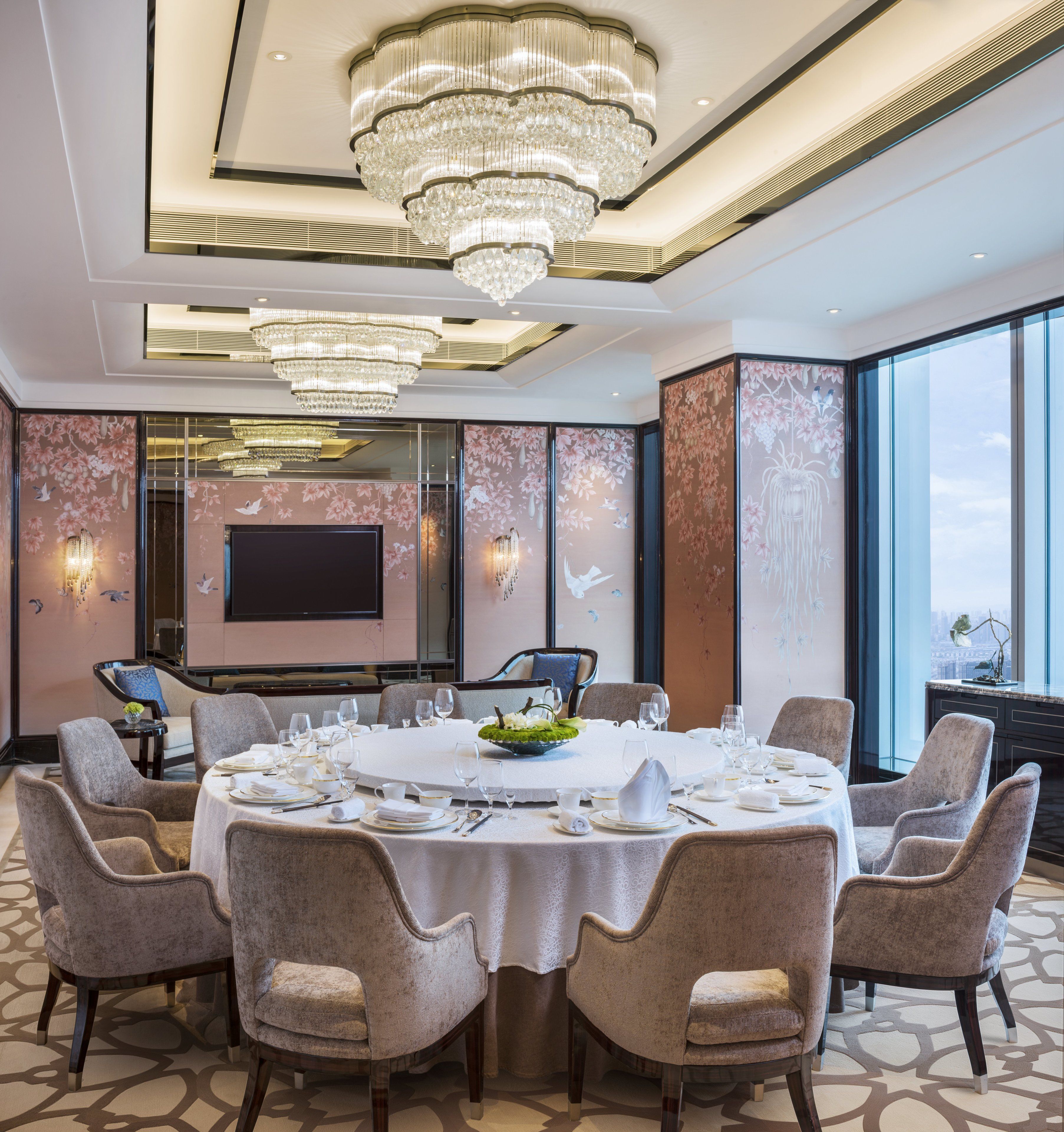 A spacious conference room with a round table, comfortable chairs, a large chandelier, and a flat-screen TV on the wall.