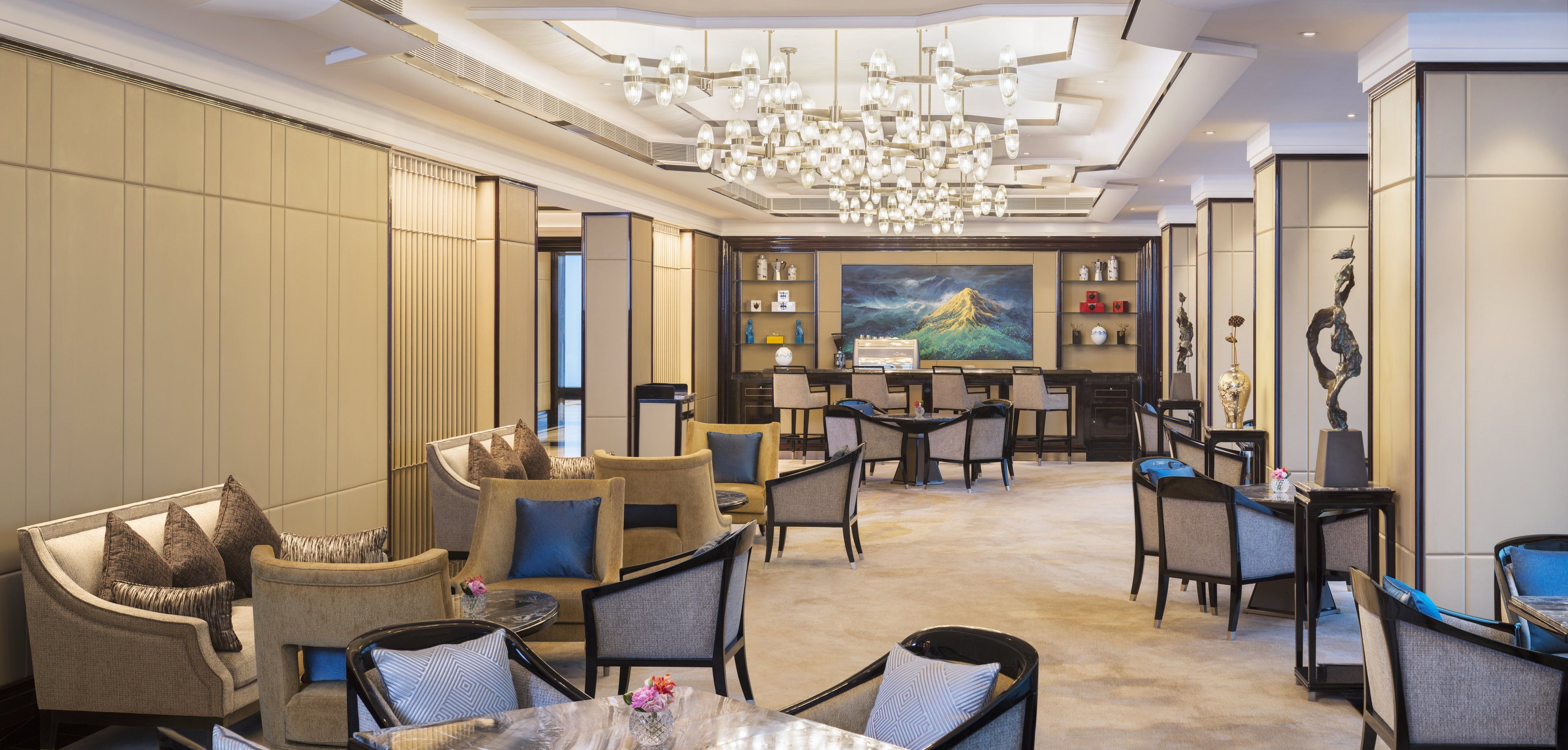 A spacious hotel lobby with a modern chandelier, comfortable seating, a large painting, and a bar area.