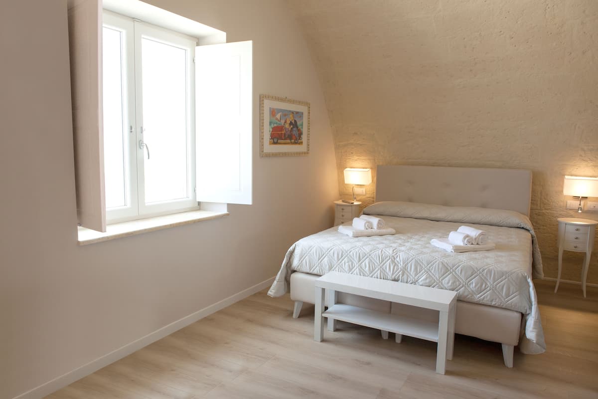 A bedroom with a bed, bedside lamps, a bench, a window, and a painting on the wall.