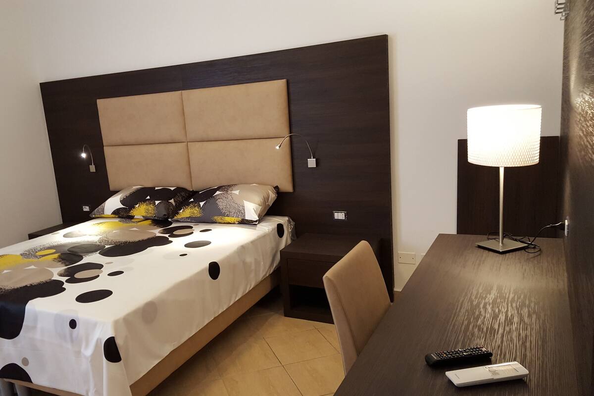 A hotel room with a bed, a bedside table, a lamp, and a remote control.