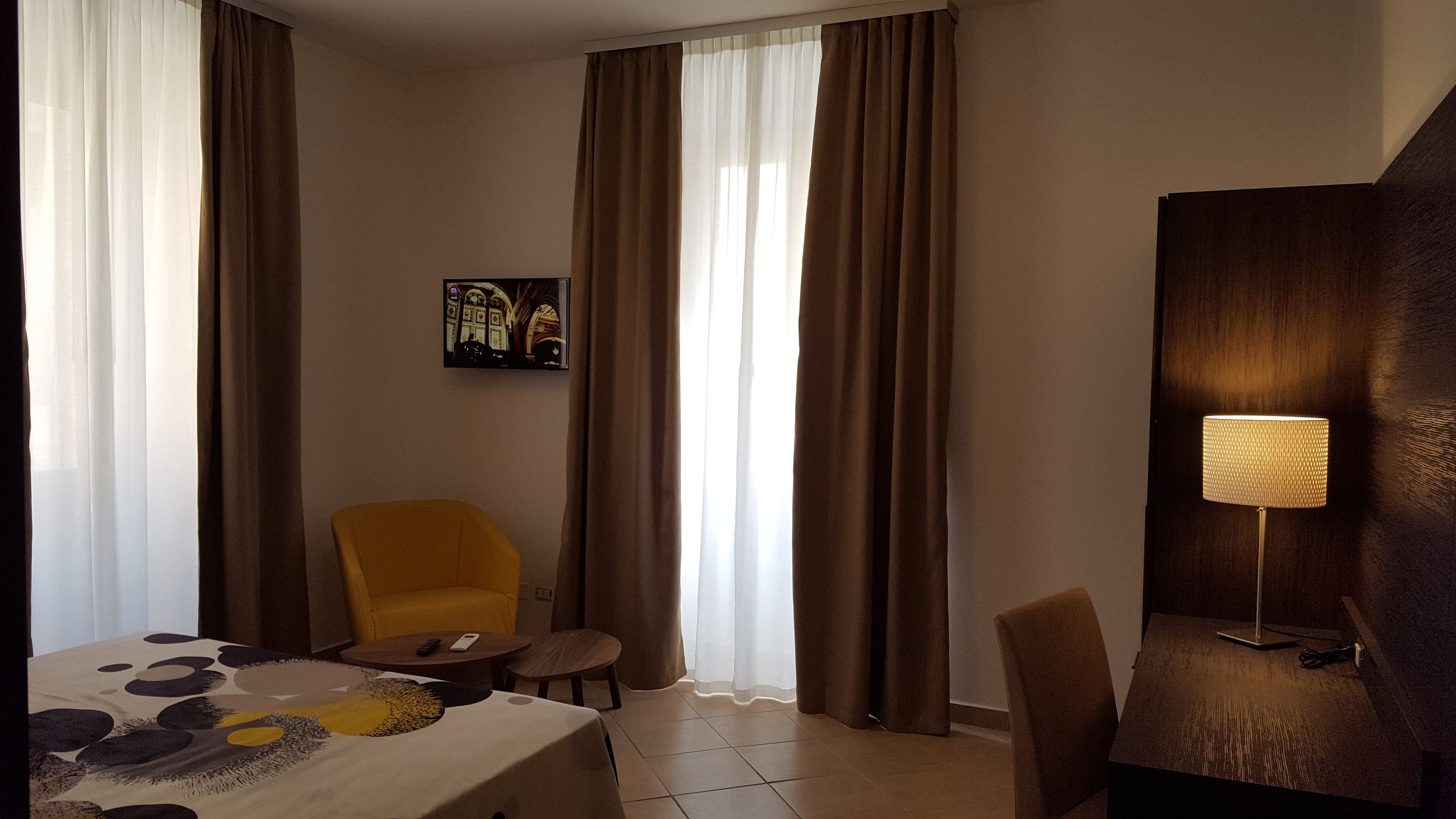 A hotel room with a bed, a yellow armchair, a small table, a lamp, and a painting on the wall.