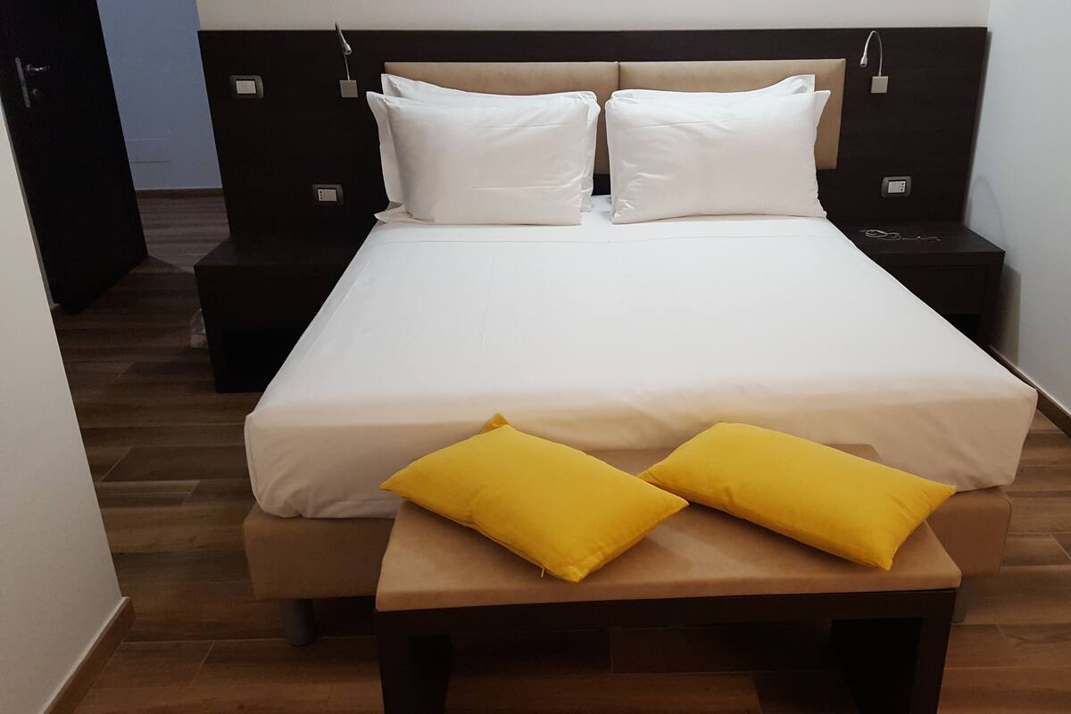 A neatly made bed with white linens and two yellow pillows, a bedside table, and a bench in front of the bed.