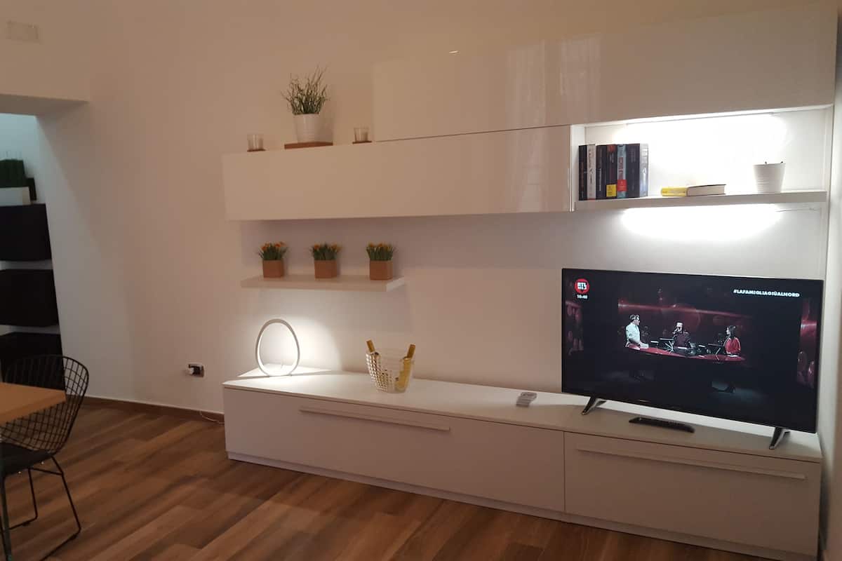 A modern living room with a TV, shelves with decorative items, a dining area with a table and chairs, and a wooden floor.