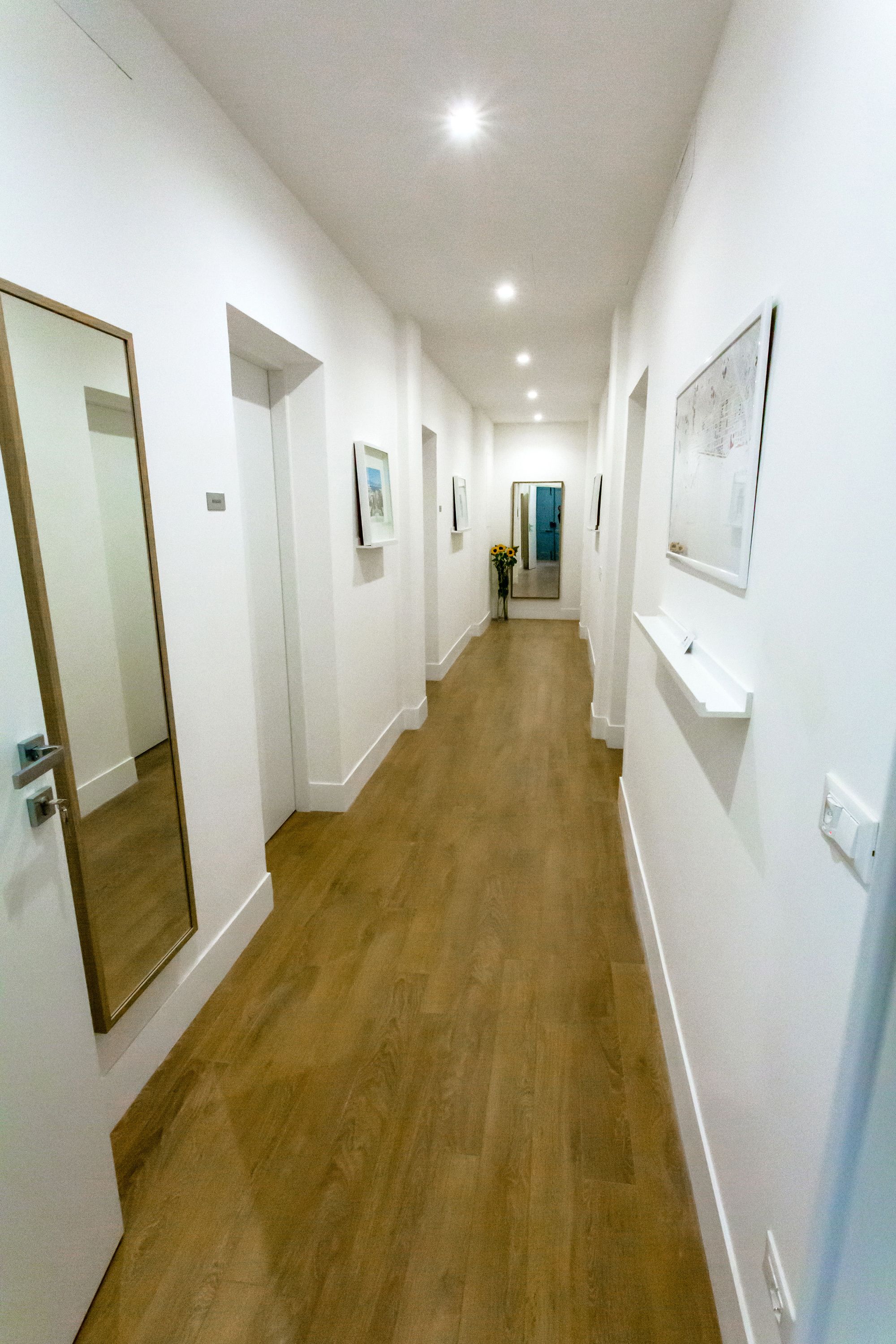A hallway with white walls, wooden flooring, and recessed lighting. There are framed pictures on the walls and a mirror on the left.